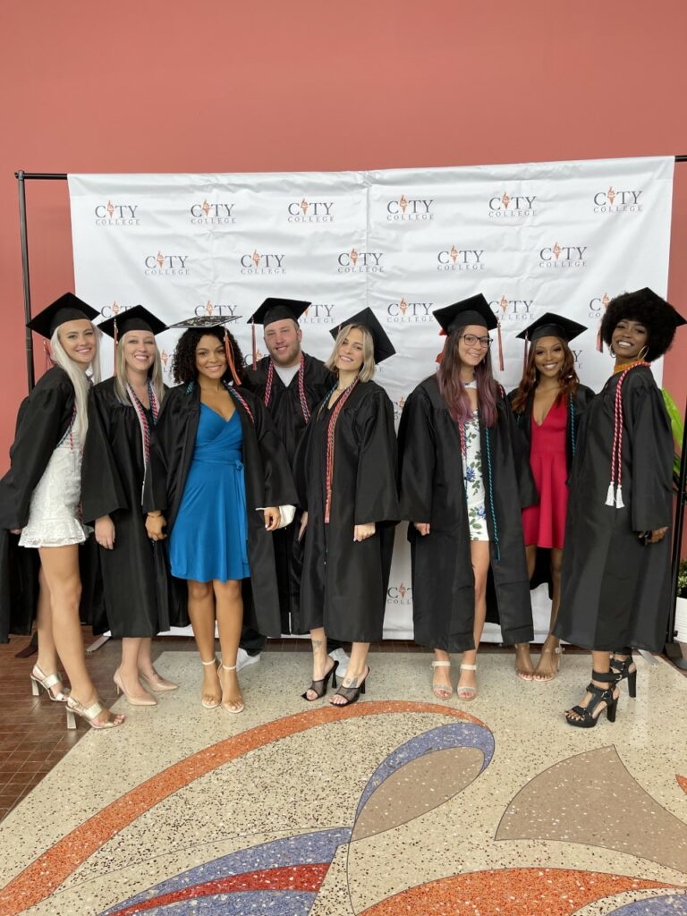 Photo from the south Florida graduation ceremony in 2021. Photo depicts students dressed in caps and gowns in front of a City College backdrop.